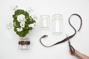 Next to a geranium and empty jars, a hand is cutting a leather belt with a pair of scissors.