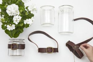 Next to a geranium and jars, a hand sews a strap to a belt, close to a buckle.