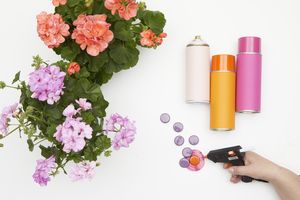 Next to two geraniums and spray cans, a hand holds a hot glue gun and glues bottle tops to a metal lid to make upcycled flower picks.