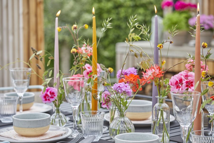 Burning taper candles and colourful cut geraniums, and grasses in glass vases on garden table