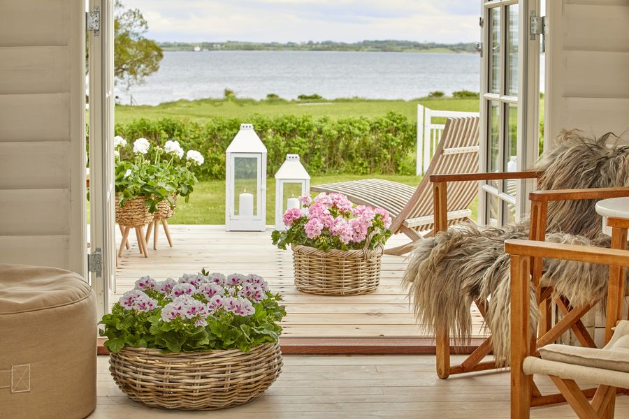 Blick von einem Raum mit Holzstühlen und Edelgeranien durch die Terrassentür auf eine Terrasse mit Liegestuhl, Gartenlaternen und Bastkörben mit Geranien