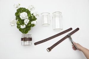 Next to a white geranium and empty glass jars, a hand punches holes in a leather belt with a hole punch.