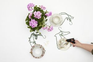 Geranium and hand holding hot glue gun, gluing cup to saucer with metal chains for DIY feeding station. Next to it are other vintage upcycled hanging decorations.