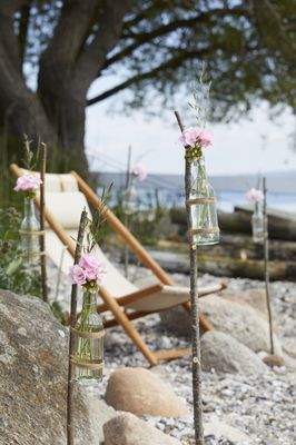 On a pebble stands a deck chair, with homemade poles made from branches to which bottles filled with geraniums and grasses have been tied