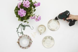 Hand holding cordless drill and drilling holes in vintage saucers next to lilac geranium