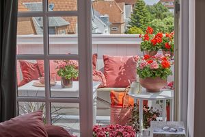 Blick durch weißes Sprossenfenster und offene Balkontür auf Stadtbalkon mit weißem Tisch, roten und apricotfarbenen Geranien, roten Kissen und  Korbtasche